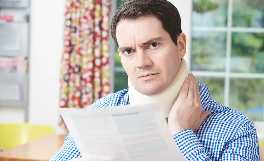 A man grimacing in pain as he holds a paper, indicating discomfort in his neck.