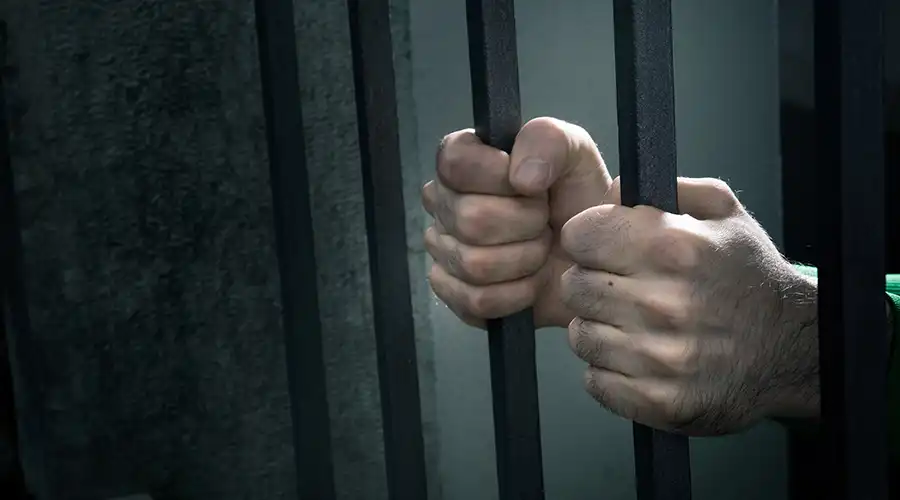 Image of a man's hands behind bars in a prison cell.