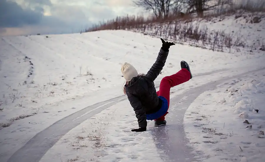A person slips and falls backwards on a snowy, icy path, legs in the air, wearing a gray jacket and a white hat, surrounded by a winter landscape.