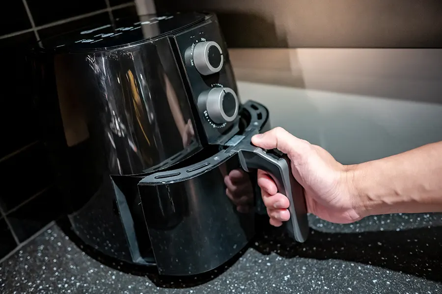 A person's hand removing a glossy black air fryer basket from the main unit positioned on a kitchen countertop.