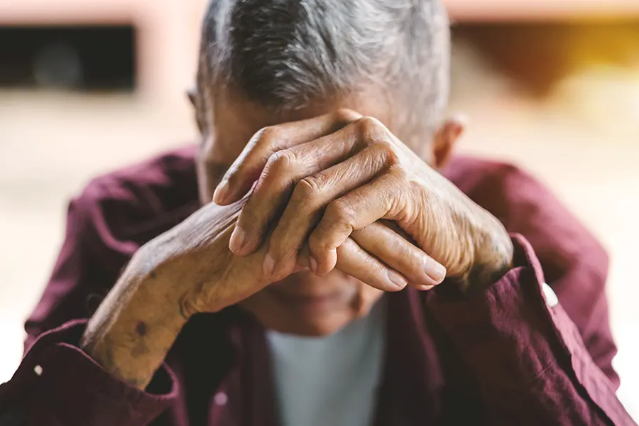 An elderly man in a burgundy shirt, sitting with his head bowed, hands clasped at the back of his neck in a posture of contemplation or fatigue.